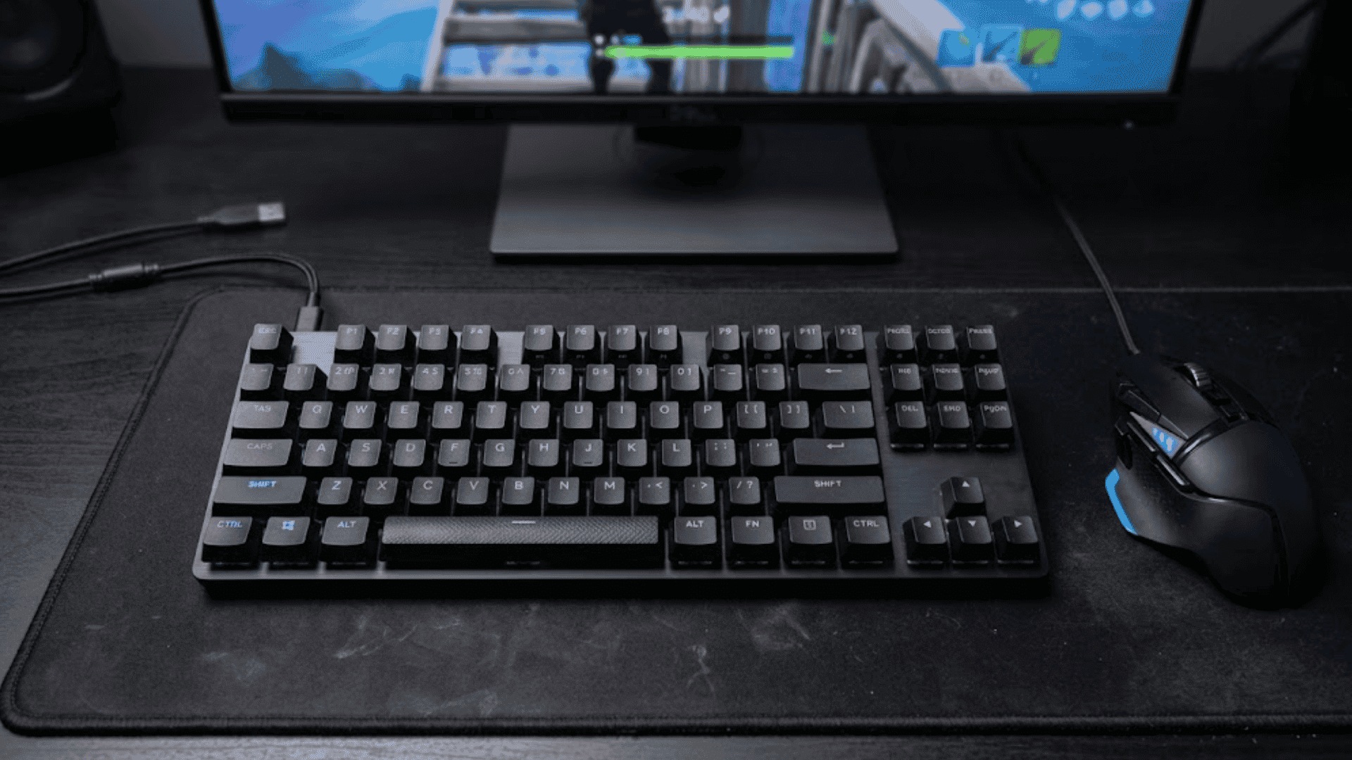A gaming keyboard and mouse on a desk, with keybinds illuminated and Fortnite gameplay on the monitor behind.