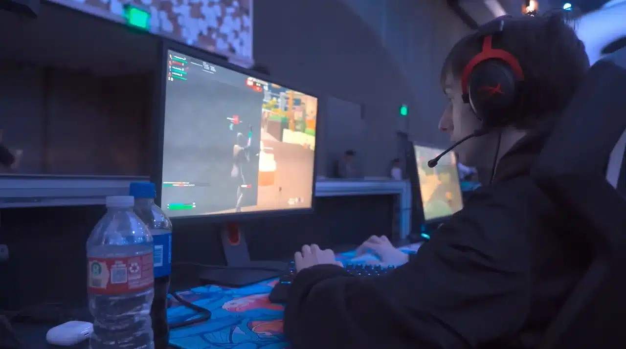 fortnite player testing competitive settings at a LAN setup wearing a HyperX headset with a custom mousepad and monitor displaying active gameplay