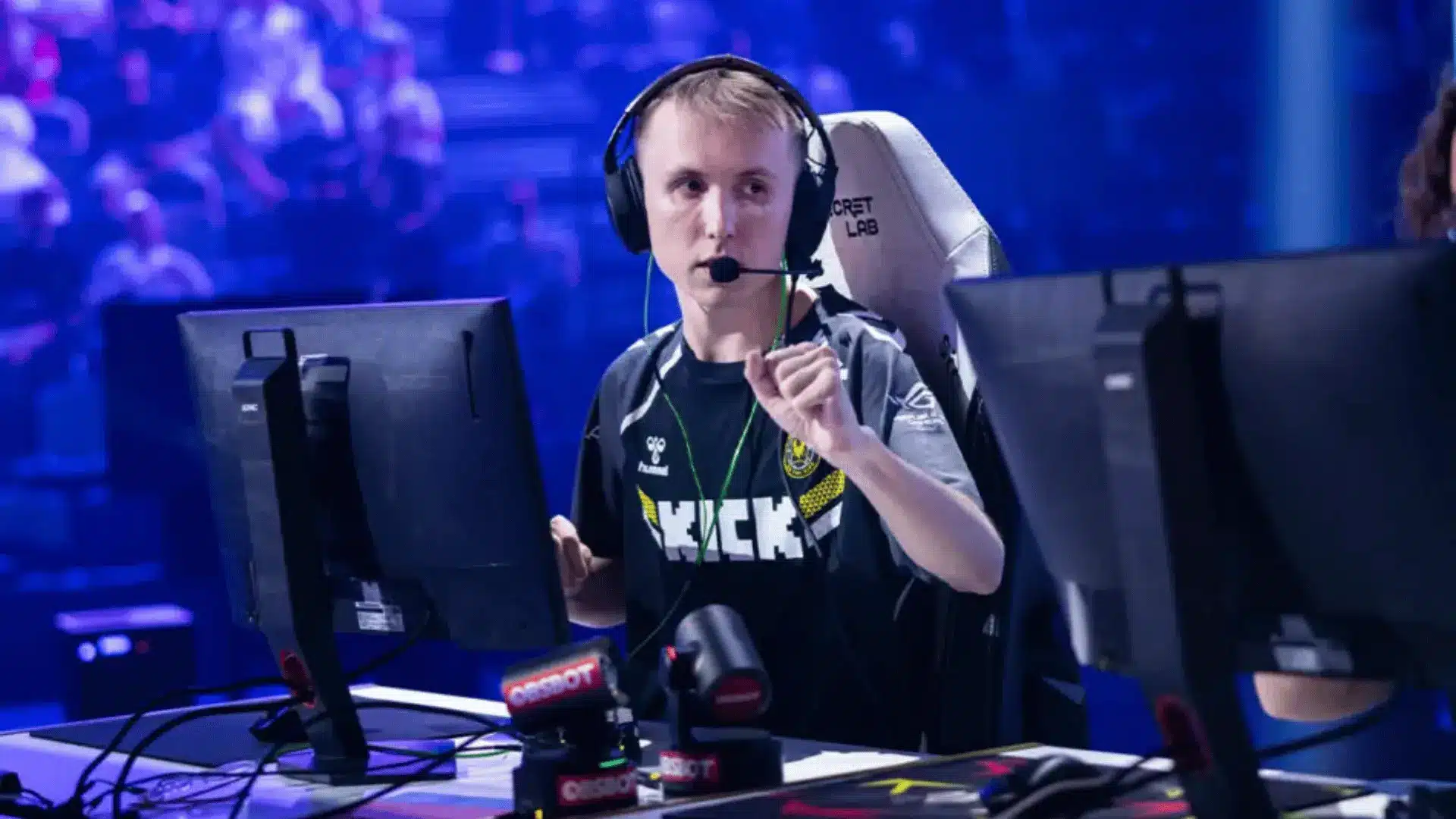 Ropz sitting at a CS2 tournament desk wearing a headset, focused on the game screen in arena background.