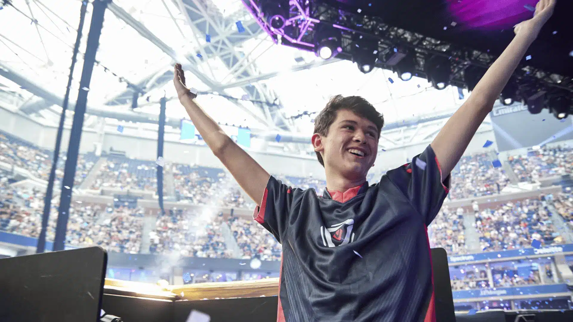 Esports player Bugha celebrates his victory with arms raised as confetti falls in a packed stadium.