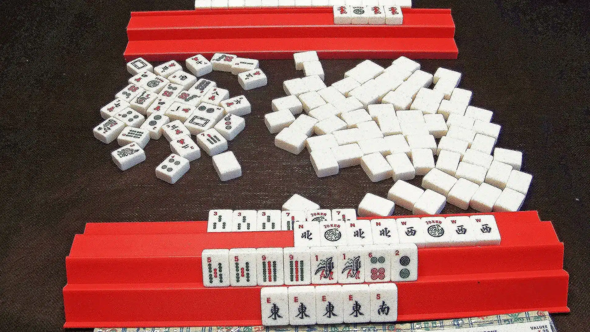 Mahjong tiles piled on a dark table between two red plastic tile racks containing organized sets.