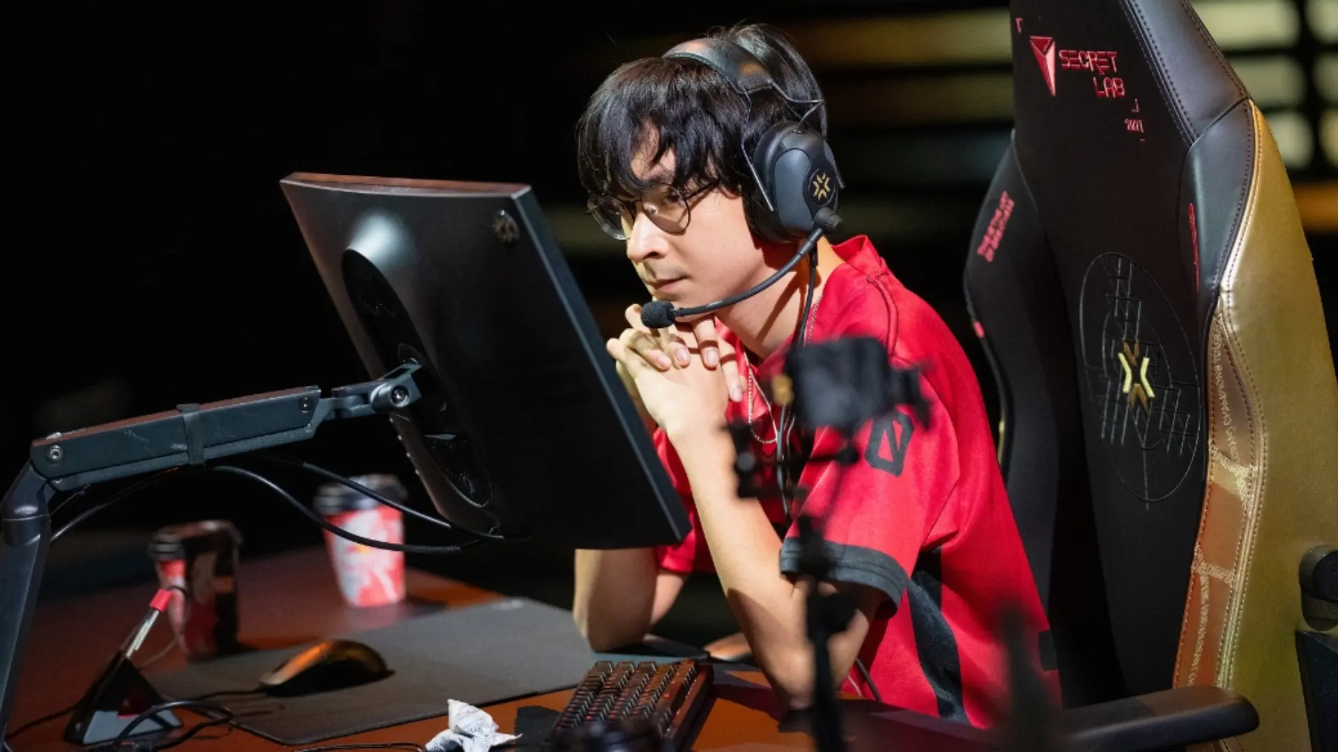 Valorant esports player wearing a red jersey and headset while focusing on a gaming setup