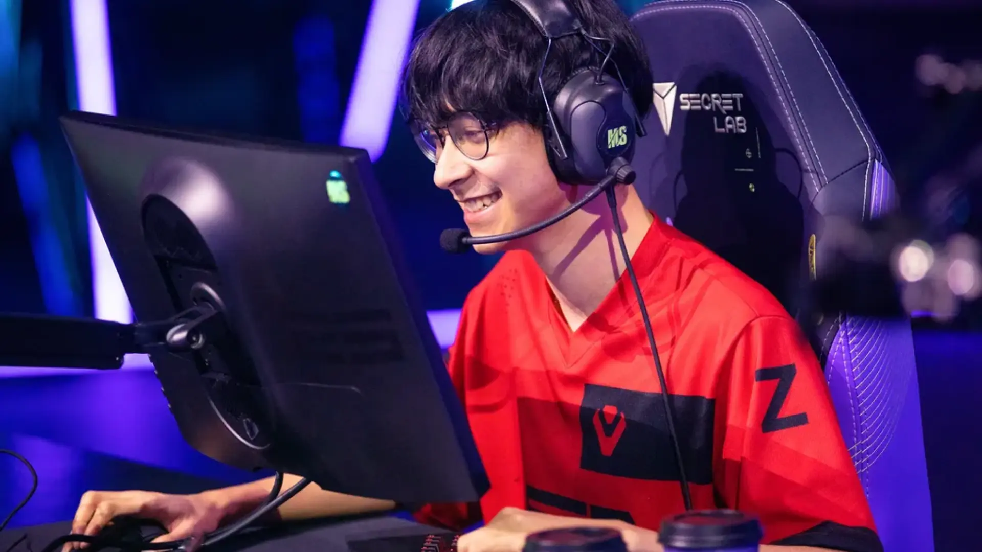 Valorant esports player wearing a red team jersey and headset while smiling at a gaming monitor