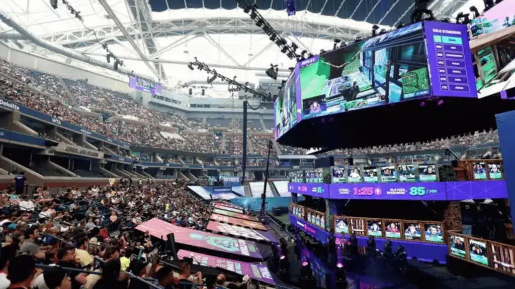 Crowded Fortnite tournament arena showing live gameplay on large screens and a packed audience watching.