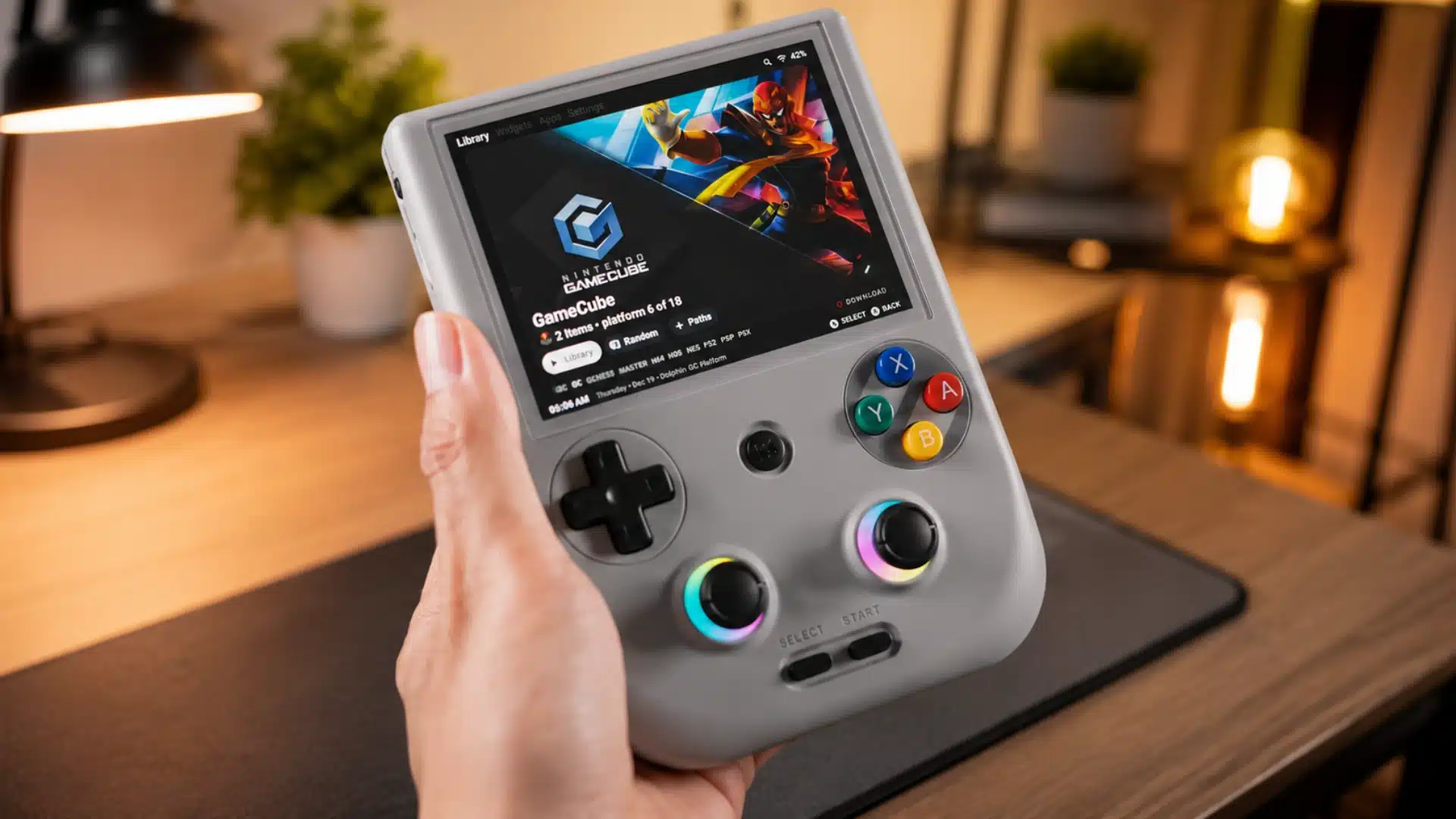 Gray handheld console displaying a GameCube library in a warm, softly lit gaming setup