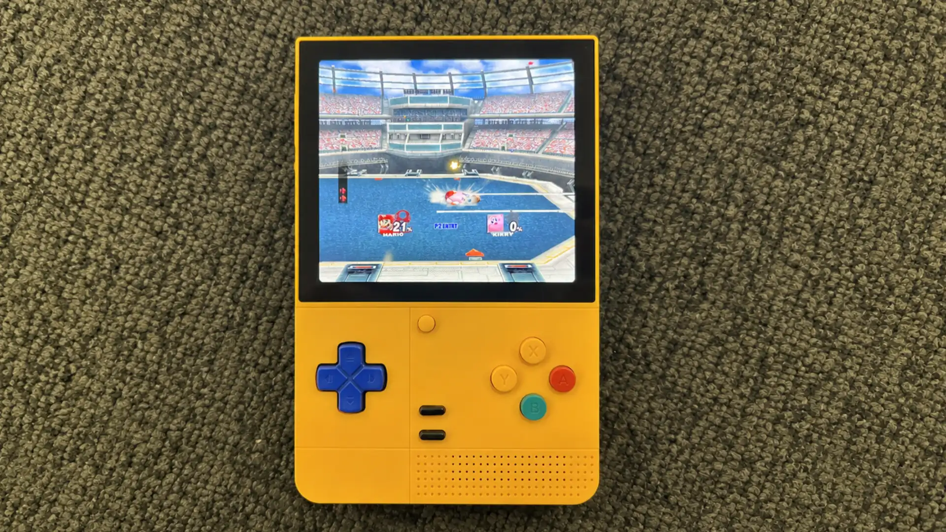 Yellow retro handheld emulator displays a fighting game while resting on textured carpet